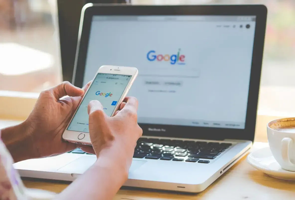 Mãos segurando um celular com a busca do Google aberta, diante de um notebook: site otimizado para o Google em Joinville